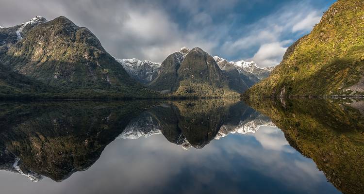 Spiegelglad fjordwater weerspiegelt feilloos scherpe, sneeuwbedekte toppen en groene valleiwanden onder een dramatische hemel.