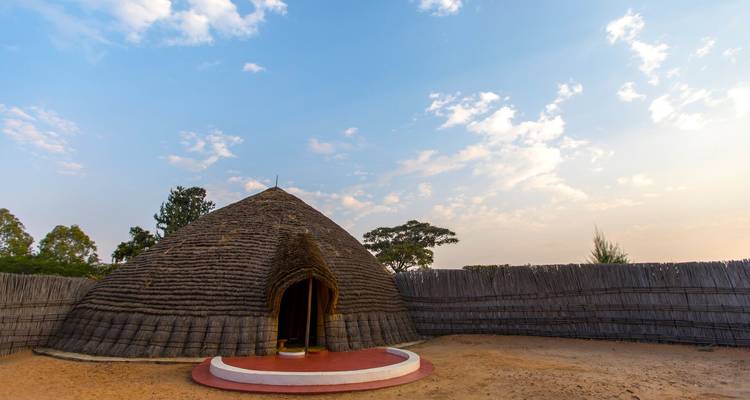 Case traditionnelle circulaire avec toit en chaume rwandais à l'intérieur d'une cour entourée d'une clôture en roseaux sous un ciel aux tons pastel.