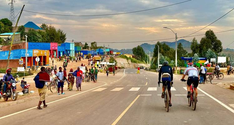 Rue animée d'une petite ville avec des cyclistes et des piétons sur fond de paysage rural montagneux.