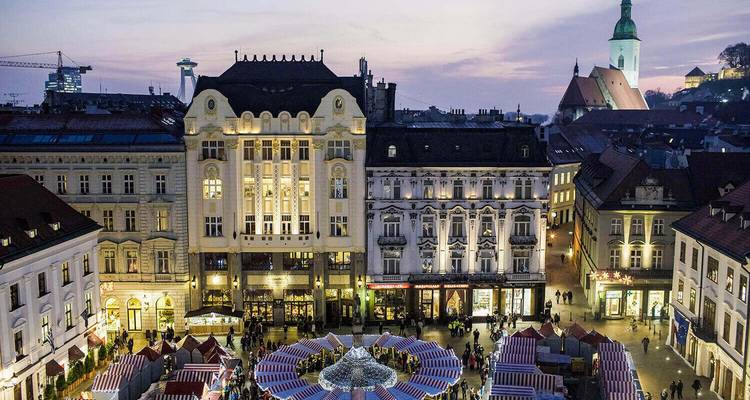 Vista nocturna de la plaza principal de Bratislava llena de mercado navideño y fachadas históricas bajo un cielo morado.