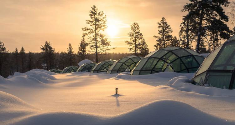 Des rangées de cabanes igloos en verre, à moitié ensevelies dans la neige profonde, brillent sous un coucher de soleil chaleureux dans une forêt de pins.