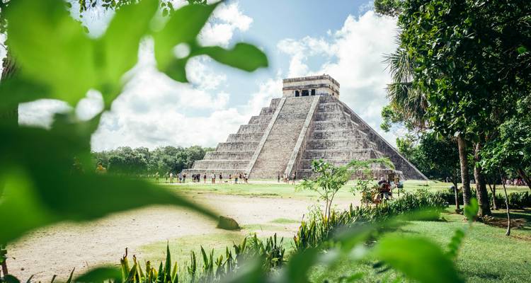 Pyramide échelonnée emblématique de Chichen Itza encadrée par un feuillage luxuriant de couleur verte sous un ciel lumineux avec quelques visiteurs à sa base.