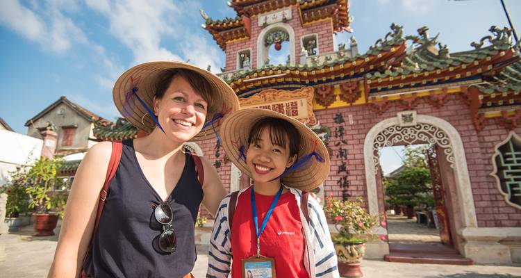 Two women in traditional conical hats stand smiling before an ornate Chinese-style gate in Hoi An.