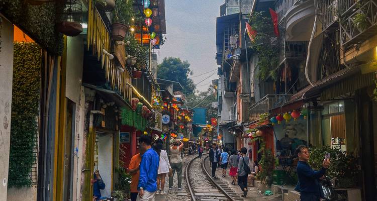 Calle estrecha y vibrante en Hanoi donde las vías del tren pasan entre casas coloridas y faroles con gente local y turistas paseando.