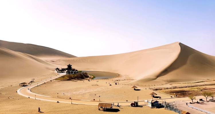 Dunas de desierto que se extienden a lo largo, con un pequeño lago oasis, un camino y construcciones de templos en el valle.