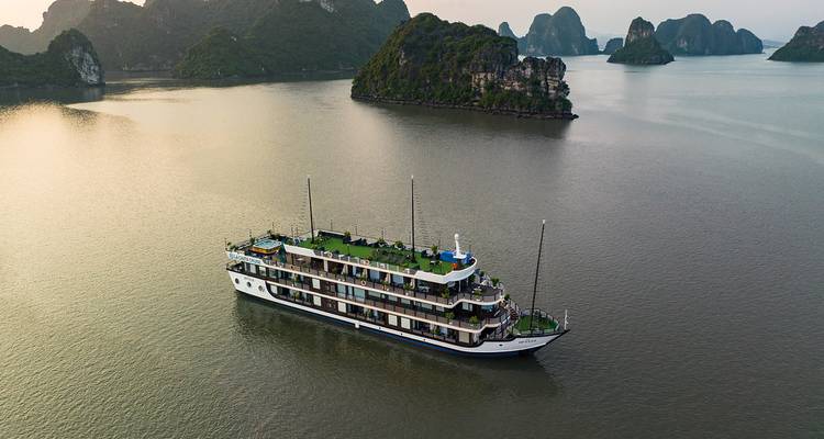 Navire de croisière de luxe naviguant dans les eaux émeraude calmes de la baie d'Ha Long parmi les îles calcaires au crépuscule.