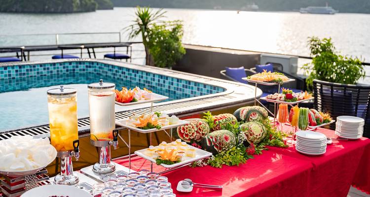 Buffet en plein air avec des fruits sculptés aux couleurs vives et des collations à côté de la piscine sur le pont de croisière donnant sur la baie.