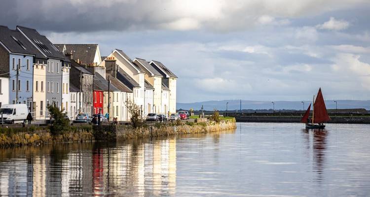 Rangée de maisons colorées en bord de mer reflétées dans une eau calme avec un petit voilier à Galway