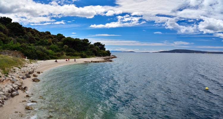 Une petite plage de galets incurvée rencontre les eaux claires et bleues de la mer Adriatique avec des visiteurs lointains se détendant sous un ciel lumineux.