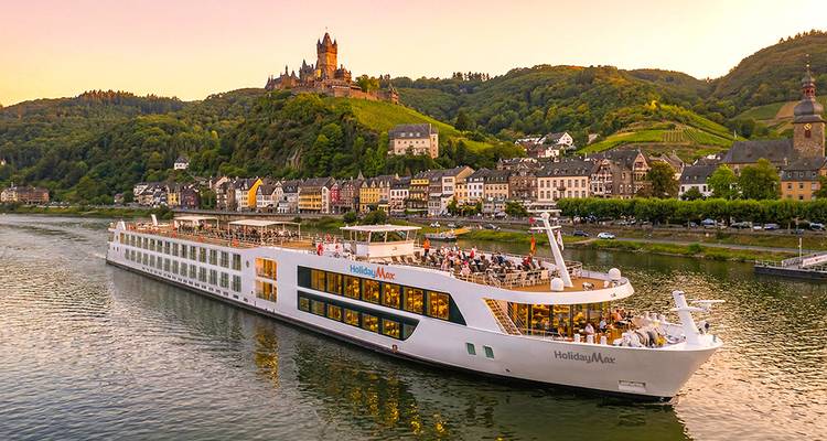 Ein elegantes weißes Flusskreuzfahrtschiff fährt bei Sonnenuntergang an der auf einem Hügel gelegenen Reichsburg Cochem und der farbenfrohen Stadt Cochem vorbei.