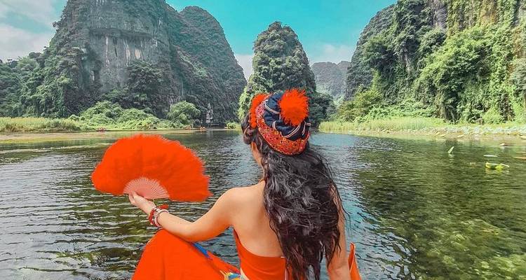 Femme vêtue d'une robe rouge éclatante tenant un éventail assise dans un bateau au milieu de pics karstiques imposants et d'une eau cristalline
