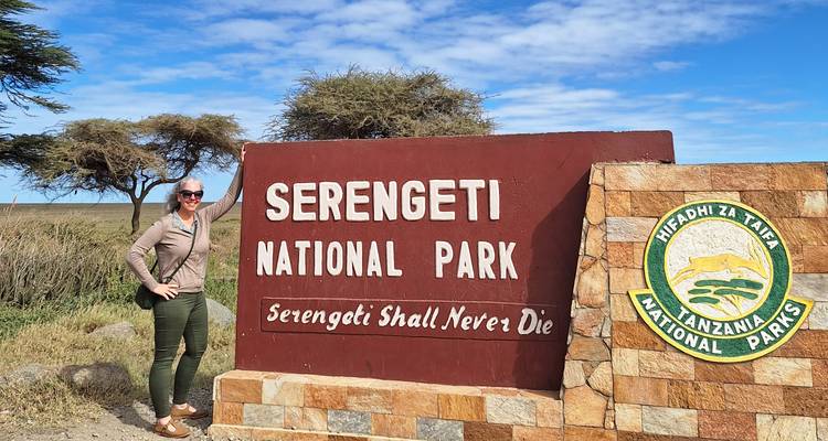 Voyageur souriant posant à côté de l'enseigne d'entrée du Parc national du Serengeti par une journée ensoleillée.