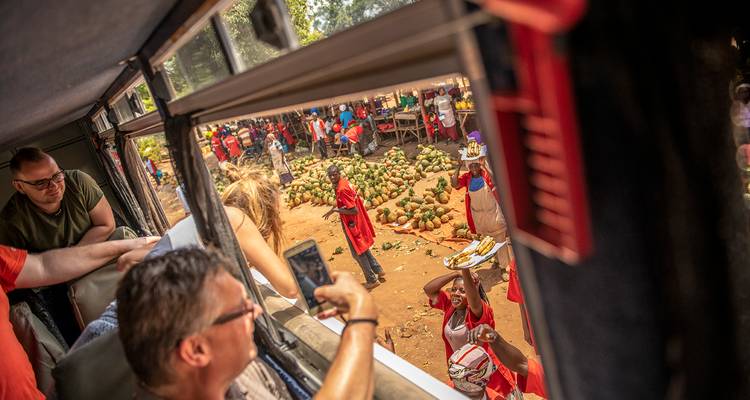 Vue depuis la fenêtre d'un autobus d'un marché de fruits routier animé avec des passagers qui photographient la scène.