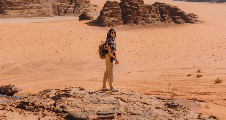 Femme voyageuse sac à dos debout sur une corniche rocheuse surplombant les immenses dunes rouges du désert de Wadi Rum.