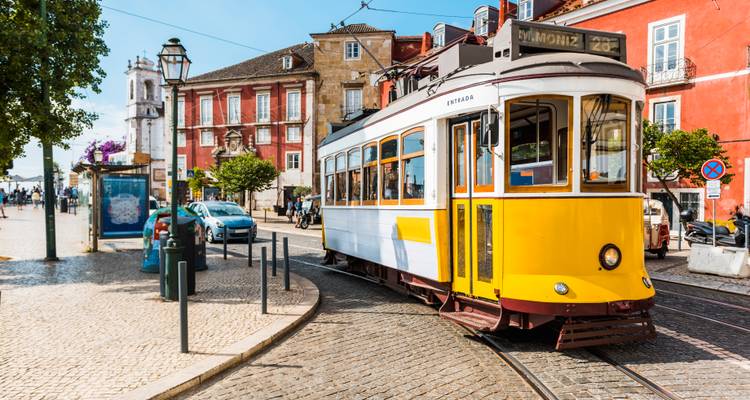 Klassische gelbe Straßenbahn, die auf eine Kopfsteinpflaster-Straße abbiegt, die von farbenfrohen historischen Gebäuden in Lissabon gesäumt ist