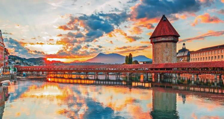 Le pont de la Chapelle emblématique et sa tour d'eau enjambant la rivière Reuss avec un ciel de coucher de soleil coloré et les montagnes reflétées dans l'eau.