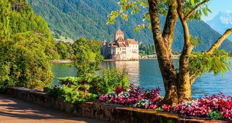 Le Château de Chillon en pierre sur un lac étincelant encadré par des fleurs en fleur et des pentes boisées.