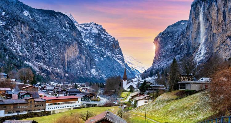 Charmant village alpin niché entre des falaises enneigées escarpées sous un ciel de coucher de soleil aux teintes pastel.
