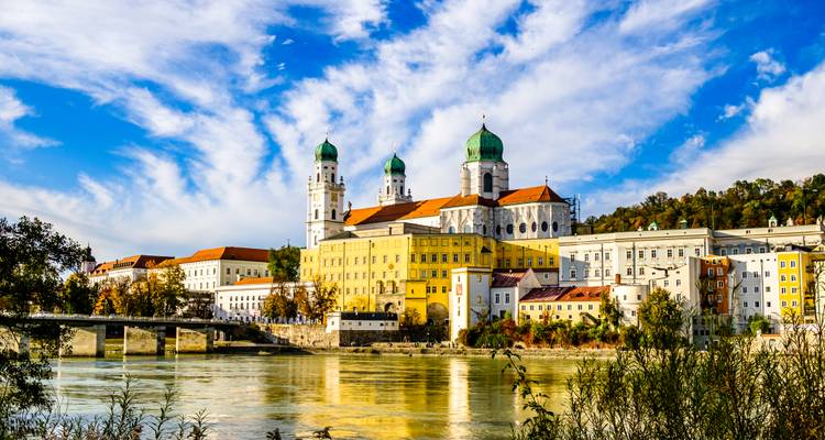 Der Stephansdom von Passau mit seinen grünen Kuppeln erhebt sich über farbenfrohen Gebäuden am Fluss.