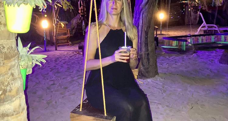 Une femme est assise sur une balançoire en bois dans un bar de plage illuminé par des lumières violettes éclatantes la nuit.