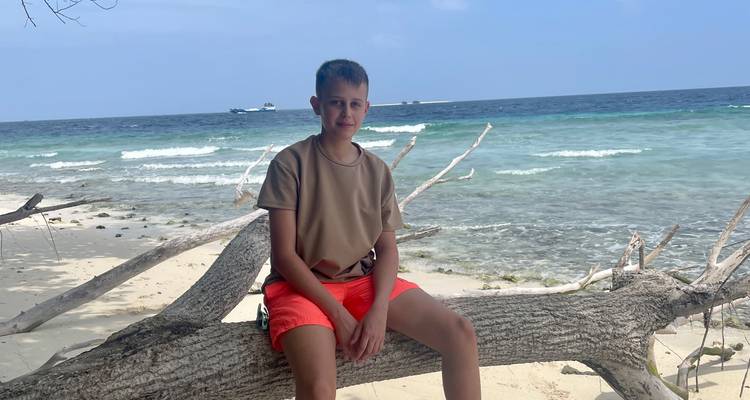 Un adolescent assis sur un tronc d'arbre tombé sur une plage sauvage avec des vagues turquoise derrière lui.