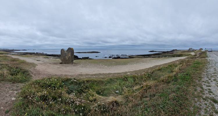 Large panorama côtière avec une pierre levée isolée (menhir) près du rivage sous un ciel nuageux.