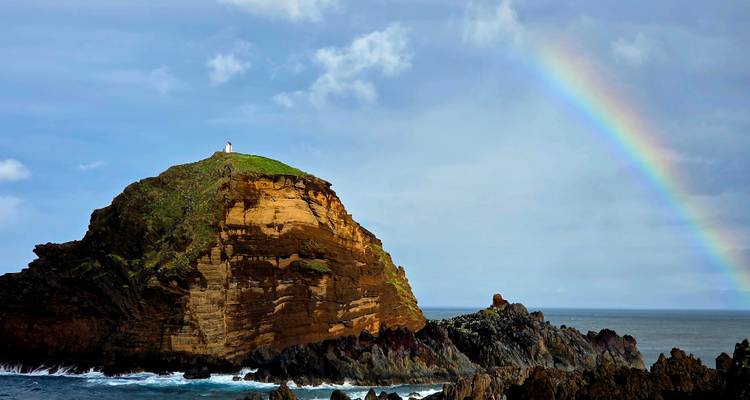 Un îlot rocheux surmonté d'un phare rencontre la mer tandis qu'un arc-en-ciel éclatant s'élève au-dessus.