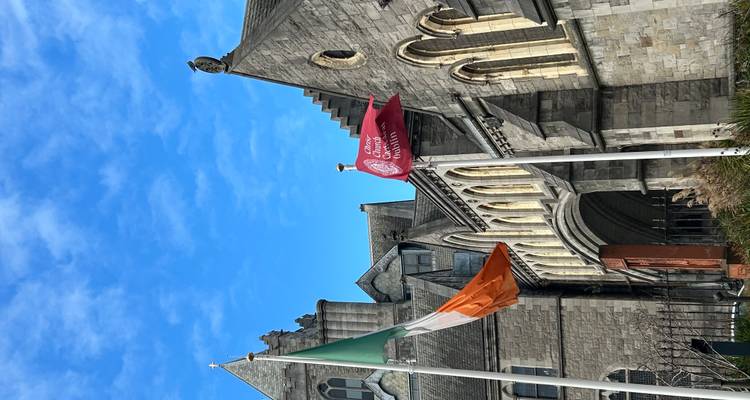 Irish tricolour and a red flag waving in front of a stone church facade under a vivid blue sky.