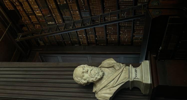 Marble bust of Socrates beside towering book stacks and a tall ladder in a dark wooden library.