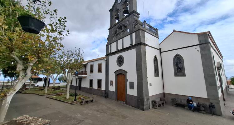 Église en pierre avec clocher par un jour nuageux, une personne seule assise sur un banc à l'extérieur.