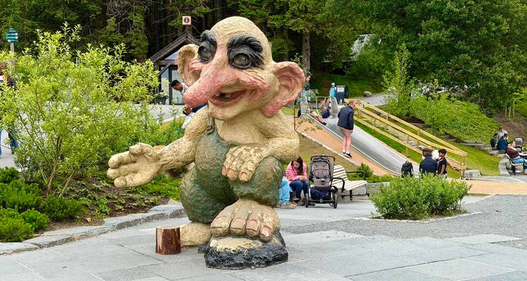 Grande statue de troll fantaisiste dans une zone de parc avec des enfants et des parents à proximité et une aire de jeux en arrière-plan.