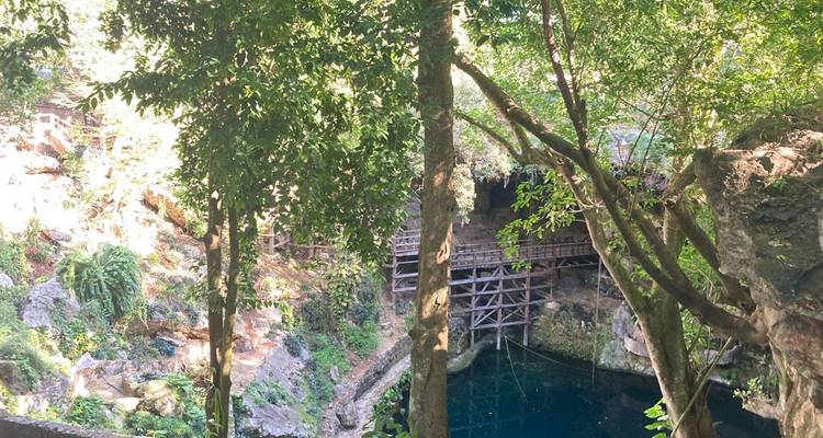 Une jungle luxuriante entoure un cenote bleu profond accessible par des escaliers et des plateformes en bois.