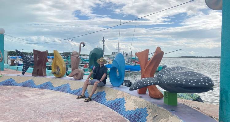 Voyageur assis près des grandes lettres colorées du port de HOLBOX avec des bateaux de pêche en arrière-plan