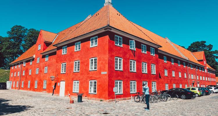 Edificio de estilo cuartel de color rojo brillante con techo de tejas naranjas sobre un cielo azul despejado, turista parado al frente.