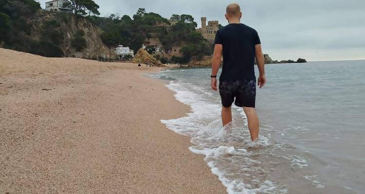 Un homme marchant pieds nus dans le ressac peu profond le long d'une plage de sable avec un château perché sur une falaise en arrière-plan.