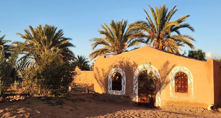 Pequeña casa de huéspedes de adobe con puertas pintadas artísticamente rodeada de palmeras bajo la luz del atardecer.