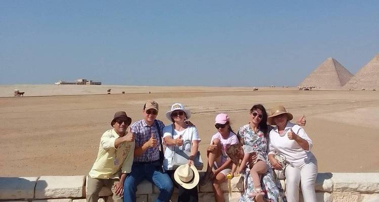 Des touristes levant le pouce tandis qu'ils sont assis sur un muret bas, les pyramides de Giza lointaines émergeant du désert derrière eux.
