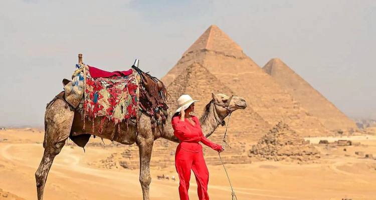 Femme vêtue d'une tenue rouge frappante tenant la bride d'un chameau avec deux pyramides se dressant majestueusement en arrière-plan dans le désert brumeux.
