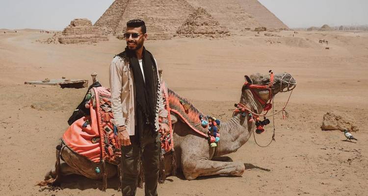 Voyageur se tenant avec élégance à côté d'un chameau reposant, richement orné, avec une pyramide s'élevant derrière eux.