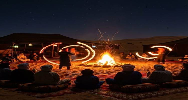 Campamento desértico nocturno donde los viajeros se sientan en tapetes alrededor de una fogata mientras los artistas hacen girar poi de fuego bajo las estrellas.