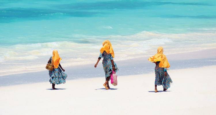 Drie vrouwen in gedessineerde jurken en oranje hoofddoeken wandelen langs een ongerepte wit zandstrand en turquoise zee.