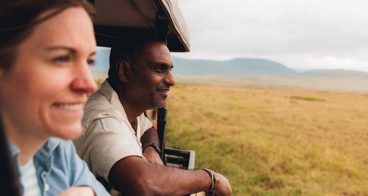Safari-gangers leunen uit een jeep en genieten van het uitzicht over open graslanden onder een bewolkte lucht.