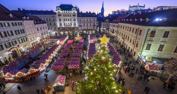 Vue aérienne sur un sapin de Noël brillamment illuminé et des étals de marché aux motifs colorés dans la vieille ville de Bratislava avec le château en arrière-plan.