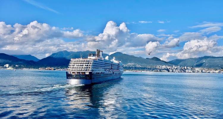 Large cruise ship departing Vancouver harbor with coastal mountains and city skyline under bright blue skies.