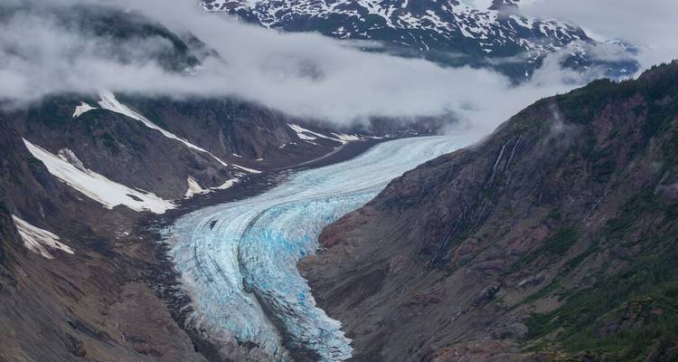 Ein massiver Gletscher windet sich durch ein raues Bergtal, das teilweise in neblige Wolken gehüllt ist.