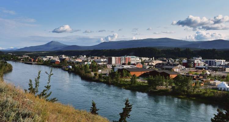 Flusslandschaft mit niedrigen Gebäuden vor hügeliger Landschaft unter blauem Himmel.