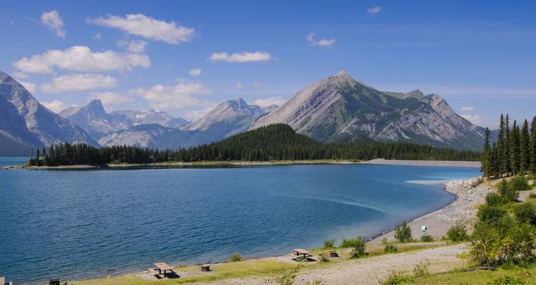 Hellblauer Alpensee umgeben von Picknicktischen, Kiefernwald und zerklüfteten Rocky Mountains.