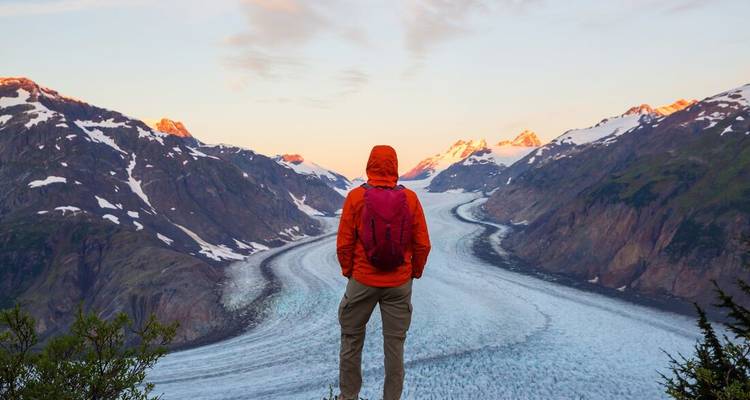 Ein Wanderer in einer orangefarbenen Jacke steht auf einem felsigen Vorsprung und blickt auf einen ausgedehnten, sich windenden Gletscher, der von schneebedeckten Bergen in warmem Abendlicht flankiert wird.