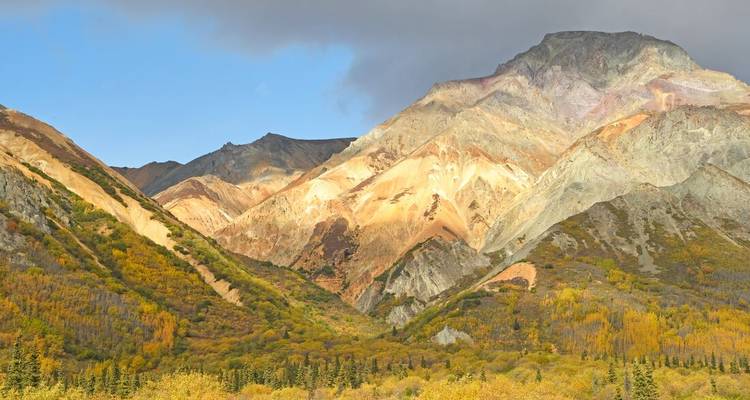 Zerklüftete mehrfarbige Berge ragen über Herbstwäldern unter einem teilweise bewölkten blauen Himmel auf.