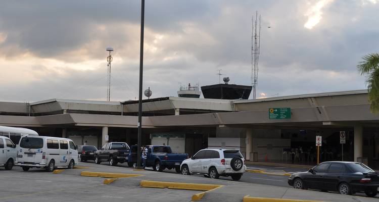 Extérieur d'un petit terminal d'aéroport tropical avec des voitures garées sous un ciel nuageux